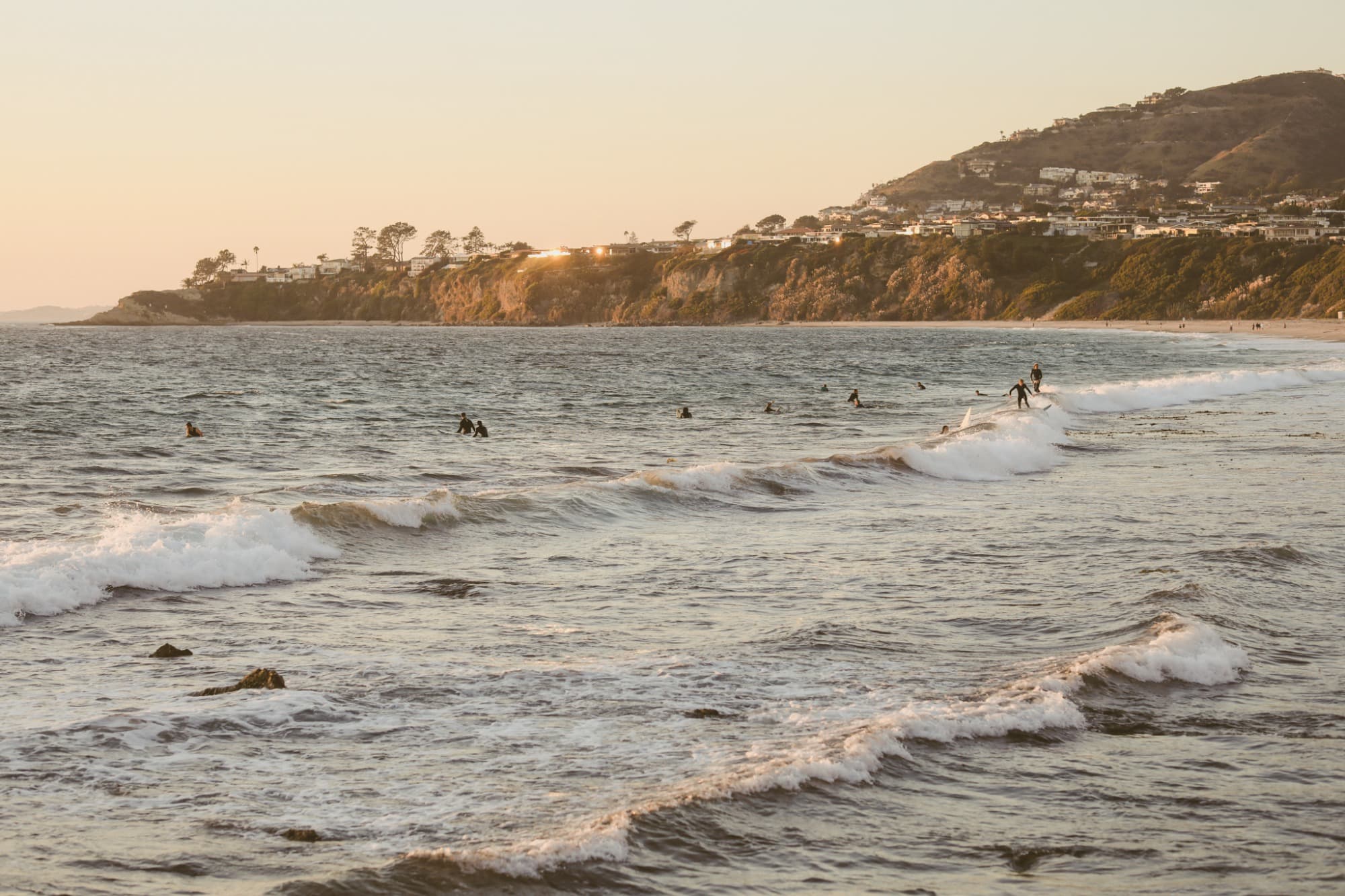 Surfers in ocean waves beneath a coastal hillside at sunset